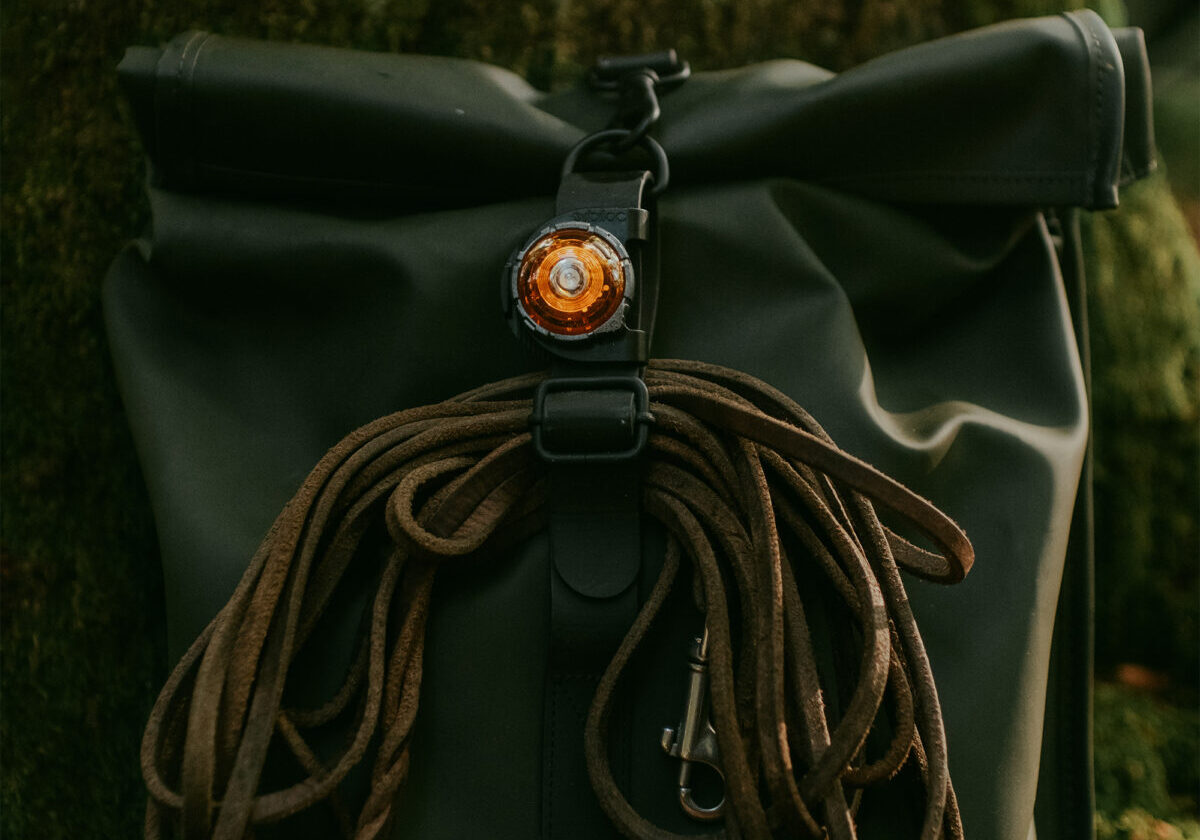 "Close-up van de bovenkant van een rugzak met opgerolde lange leren leiband en amberkleurige Orbiloc Safety Light en Clip