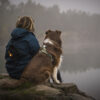 Persoon met dreadlocks en zijn Australian Shepherd zitten samen aan de oever van een mistig meer, met Orbiloc lampjes aan hun tas en halsband.
