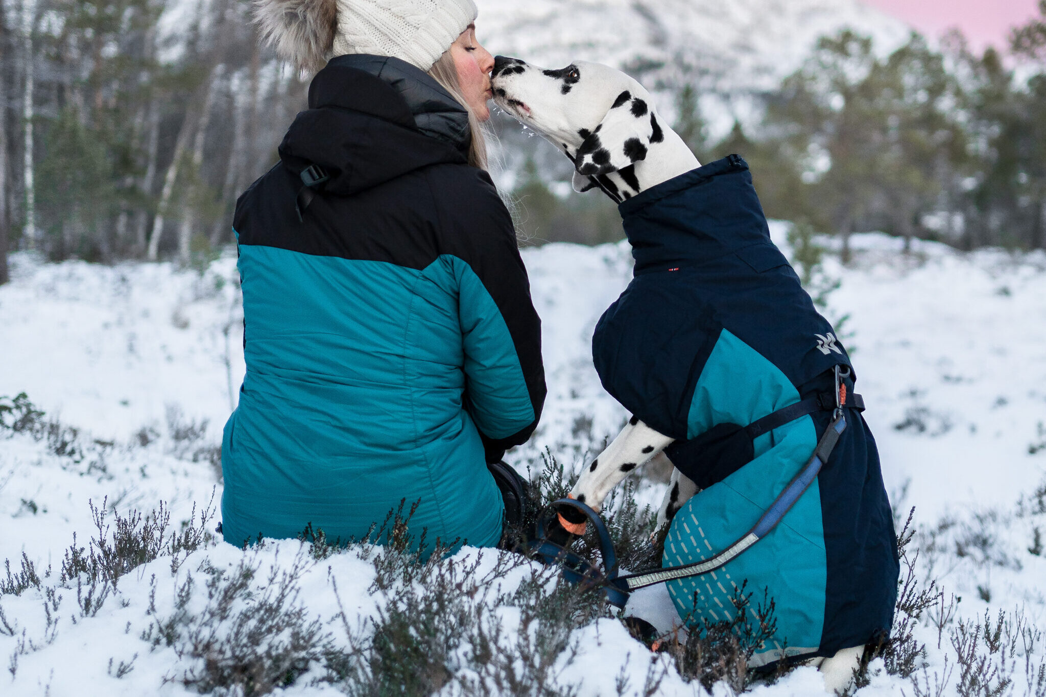 Vrouw met hond in de sneeuw in de bergen, elk met een blauwe winterjas, ze geven een kusje aan elkaar.